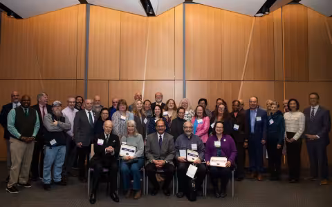 A group of smiling people, first row seated, second two rows standing, wear name tags and hold certificates.