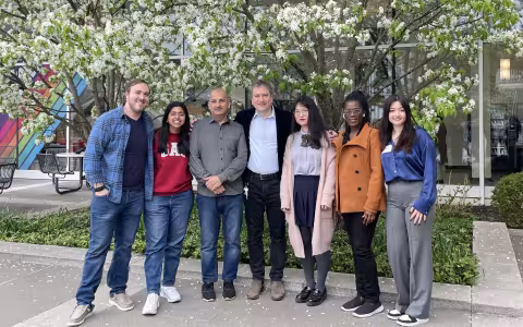 Seven people come together for a group photo outdoors. The group is standing in front of trees with white blossoms and the glass wall of UAlbany's lecture center concourse is behind them.