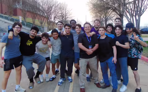 Members of the UAlbany wrestling team, including 15 young men, pose for a team photo outside of an arena in a roadway.