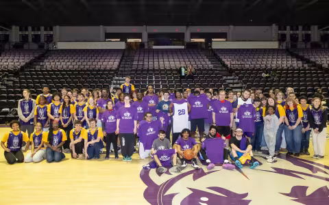 A large group of people wearing shirts with UAlbany written in all capital letters and a graphic of a Great Dane dog pose together on a basketball court with rows of empty seats behind them.