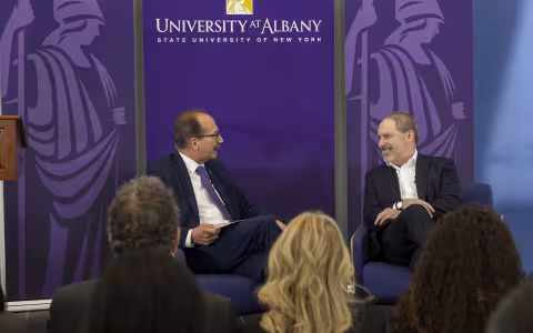 Two men are seated before a crowd of people in front of a purple backdrop with "University at Albany State University of New York" written on it with two Minerva logos adorning the sides.