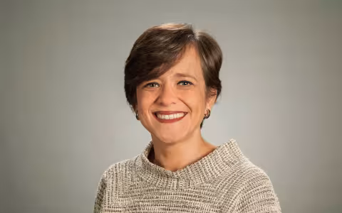 A portrait of a smiling woman with short brown hair wearing a light beige sweater against a light brown backdrop.