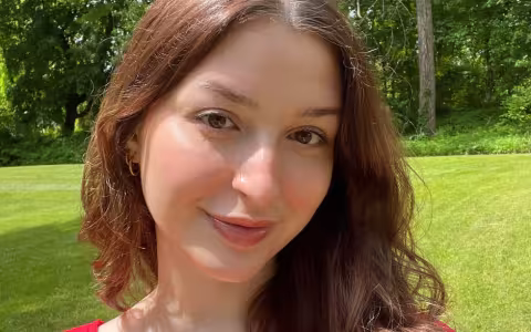 A woman with long brown hair and a red top stands outdoors on a sunny day, with green grass and trees in the background.
