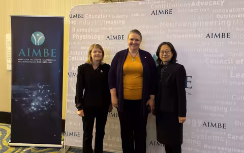 Three women smile for a photo, standing against a backdrop printed with the name “AIMBE” in purple, with a collection of science-related words in very faint print. To their left is a pull-up poster that says “AIMBE American Institute for Medical and Biological Engineering”. The woman on the left is wearing a black blazer and slacks. The woman in the center is wearing an orange top with black cardigan. The woman on the right is wearing black square rimmed glasses and a polka dot blouse under a black blazer. 