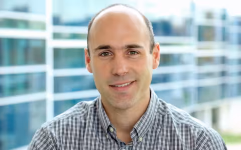 A man in a plaid shirt smiles for a portrait in front of a building with wavy glass