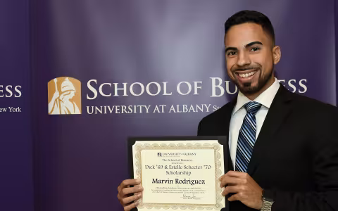 A UAlbany student with dark hair and a beard stands with an award for a Scholarship with the School of Business sign behind him.
