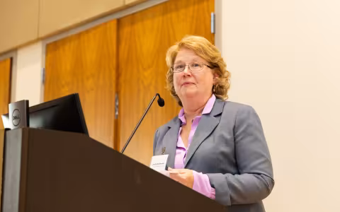 A woman with short blonde hair in a purple blouse and gray blazer stands at a podium.