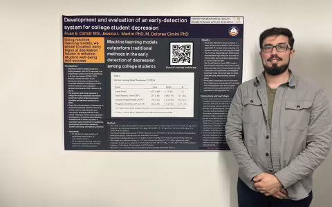 A young man with short brown hair and a goatee in black glasses, a gray button down shirt and dark pants stands against a wall next to a poster titled "Development and evaluation of an early-detection system for college student depression"