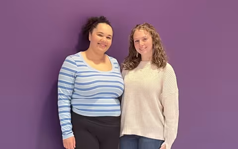 Two young women, one in a striped blue sweater and one in a beige sweater, stand smiling in front of a purple wall.