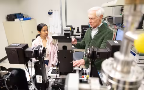 A young female student and an older male professor talk amidst numerous high-tech devices in a lab room with a filing cabinet in the background and devices in the foreground.