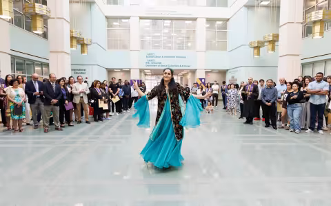 A woman wearing a black and teal dress dances inside the atrium of the Science Library while a gathered crowd watches.