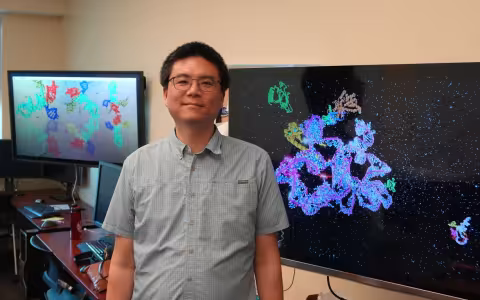 A man wearing square rimmed glasses and a green checkered button-down shirt stands in a computer lab between two large wall-mounted monitors displaying colorful visual simulations of molecules. 