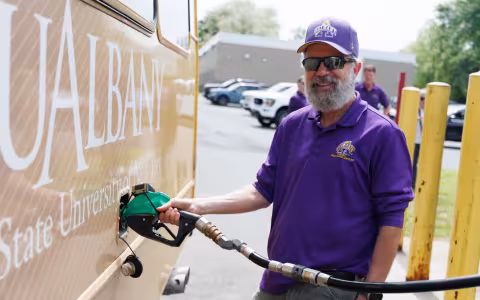 A smiling man in a purple cap and purple shirt pumps fues into a bus labeld UAlbany State University of New York