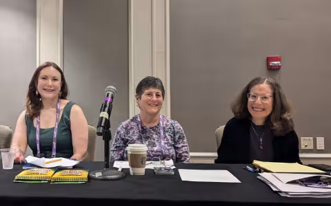 Three women sit at a conference table with a microphone, smiling warmly. Papers, notebooks, and coffee are on the table, creating a professional atmosphere.