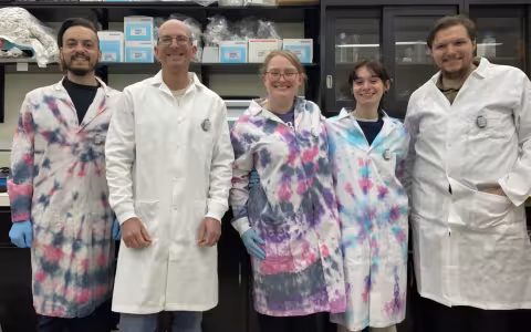 Five people, two women and three men, stand together smiling for a group portrait in front of a lab bench. All are wearing lab coats, three of which are tie dyed blue, pink and purple. Shelves of lab supplies can be seen behind them. 