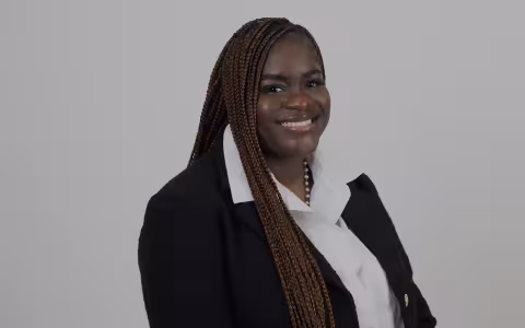 Smiling woman in a black blazer and white shirt stands against a light grey background. She has long braided hair and exudes confidence.