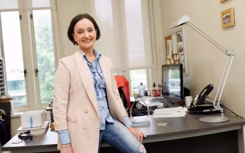 A woman with short brown hair wearing a blue blouse, cream blazer and jeans is smiling at the camera while seated on a desk.