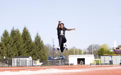 A young woman in a black T-shirt and black leggings performs a high jump at UAlbany's track and field complex on a sunny day.