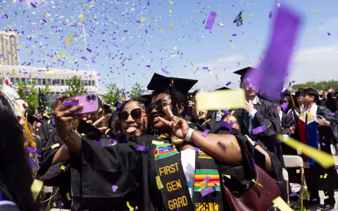Graduates take selfies with their phones as colorful confetti falls around them, celebrating their big day. 