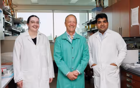 Three people, one woman and two men, stand together for a group portrait in a science lab. All are smiling and wearing lab coats.