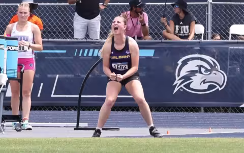 A young woman with a blond pony tail wearing a track singlet cheers while standing on the edge of the grass on a track.