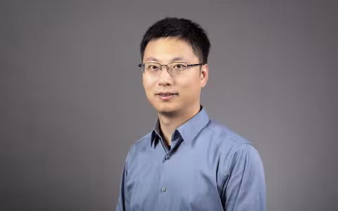 A man with short black hair and glasses in a blue shirt poses for a portrait against a gray backdrop