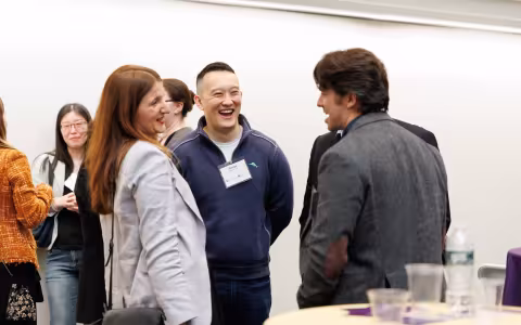 A group of people stand and talk at an indoor event; some are smiling and laughing, with name tags visible on a few attendees.
