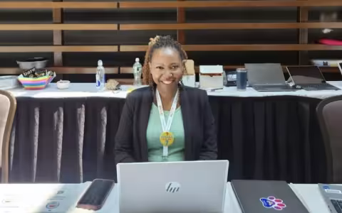 . A woman at a table uses her laptop, with various papers organized around her.