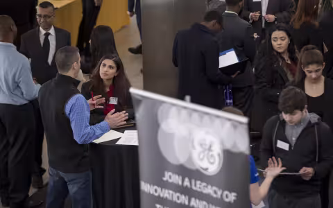 People interact at a career fair, with some standing at tables, conversing, and holding resumes. A GE banner is visible in the foreground.