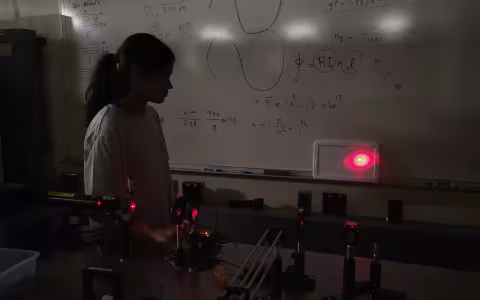 A girl with dark hair and a longsleeve shirt stands in front of a whiteboard with equations while a Raman spectroscopy laser is pointed at the wall behind her.