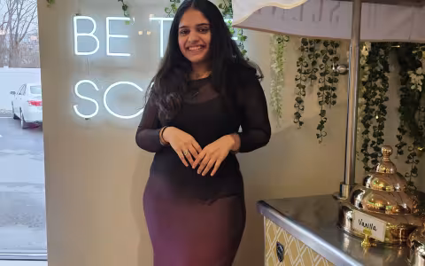 Smiling woman in a black and purple dress stands beside a vintage coffee cart. Neon sign and green vines hang on the wall, creating a cozy ambiance.