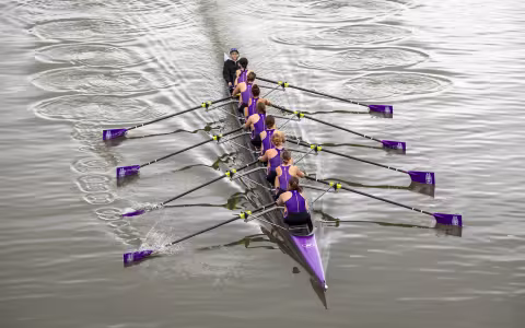 8 women and a coxswain from UAlbany row a long purple boat down a river as seen from an overhead shot
