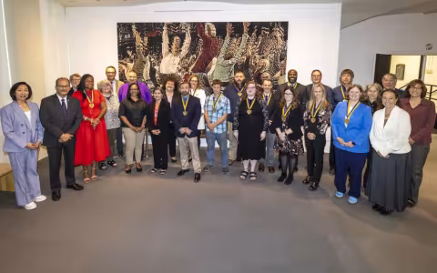 More than 2 dozen smiling people, many wearing medalions, stand in a semicircle in front of a large painting
