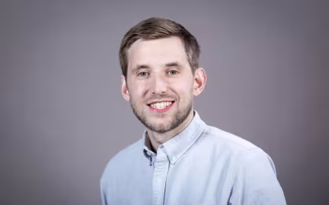A man wearing a light blue button-down shirt smiles for a portrait against a gray backdrop.