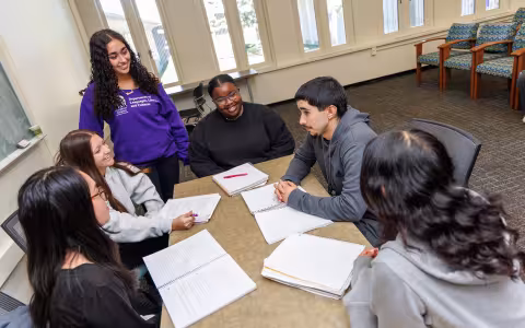 A group of students sit around a table with notebooks on a the table, one student. stands at the end of the table with a sweater that says Department of Languages, Literatures and Culture.