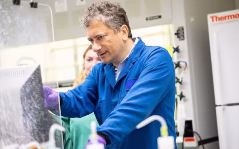 A man wearing a blue lab coat and purple gloves points to a computer screen in a brightly lit lab. 