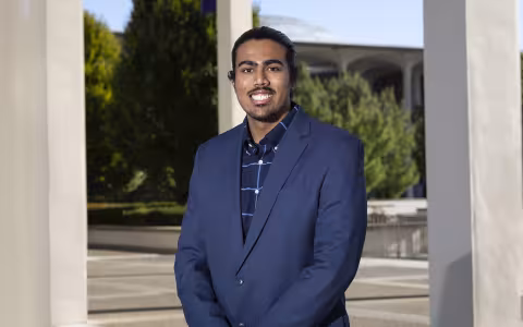A young man stands outside amid a row of columns wearing a blue blazer and a blue shirt with trees and a dome in the background outside.
