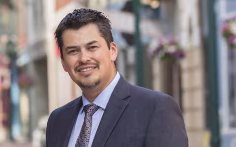 A man stands in front of a bright city street wearing a gray suit, blue shirt and gray/purple tie. He has a goatee and dark brown/black hair.