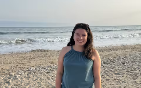 A woman stands on the beach, gazing at the ocean under a clear blue sky. 