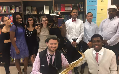 A group of people in 1920s attire pose for a photo. One is holding a saxophone and another is holding The Great Gatsby book.