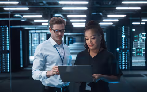Two cybersecurity professionals standing in a server room consult a laptop.