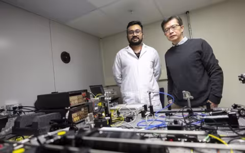 A man in a white lab coat and a man in a gray sweater stand by a table full of electrical equipment and wires