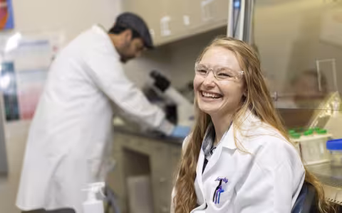 A woman with long wavy blond hair, wearing a white lab coat and clear protective goggles, smiles for a portrait in a lab. Lab benches with test tubes, and a man looking into a microscope, are in the background.
