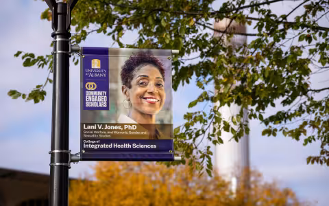 A banner hangs from a light post on campus on a sunny fall day, with the carillon in the background. The banner includes the University at Albany logo and the Community Engaged Scholars logo, as well as a the words, "Lani V. Jones, PhD, Social Welfare and Women's Gender and Sexuality Studies, College of Integrated Health Sciences" and a photo of Lani V. Jones.