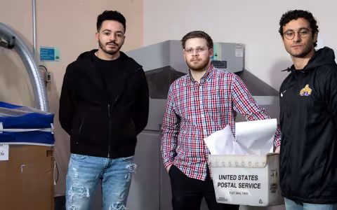 Three students stand indoors in front of a shredding machine and bypass that they created.