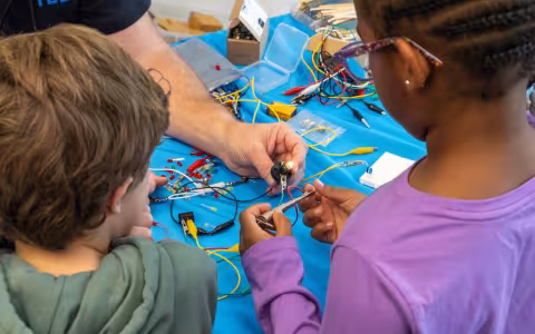 Two children stand at a table covered with wires and electronic parts as an adult helps them connect a small circuit, lighting up an LED bulb.