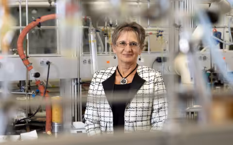 A woman with short hair, wearing glasses and a black and white tweed jacket with broad black lapels and bold accent jewelry, poses for a portrait in a chemistry lab. She is standing behind a lab bench hung with clear plastic tubing. Various tubes and glass lab containers are behind and around her. 