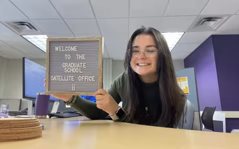 A woman holds a sign that says "Welcome to the Office," inviting guests into the workspace with a friendly gesture.