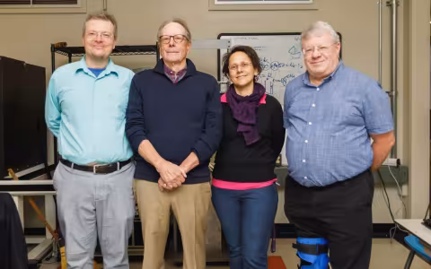 three men and a woman stand in a row facing the camera in front of a whiteboard scrawled with figures and shapes