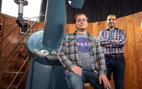 UAlbany PhD student John Bradburn sits by a large telescope in an observatory as Assistant Professor Mustafa Aksoy stands in the background